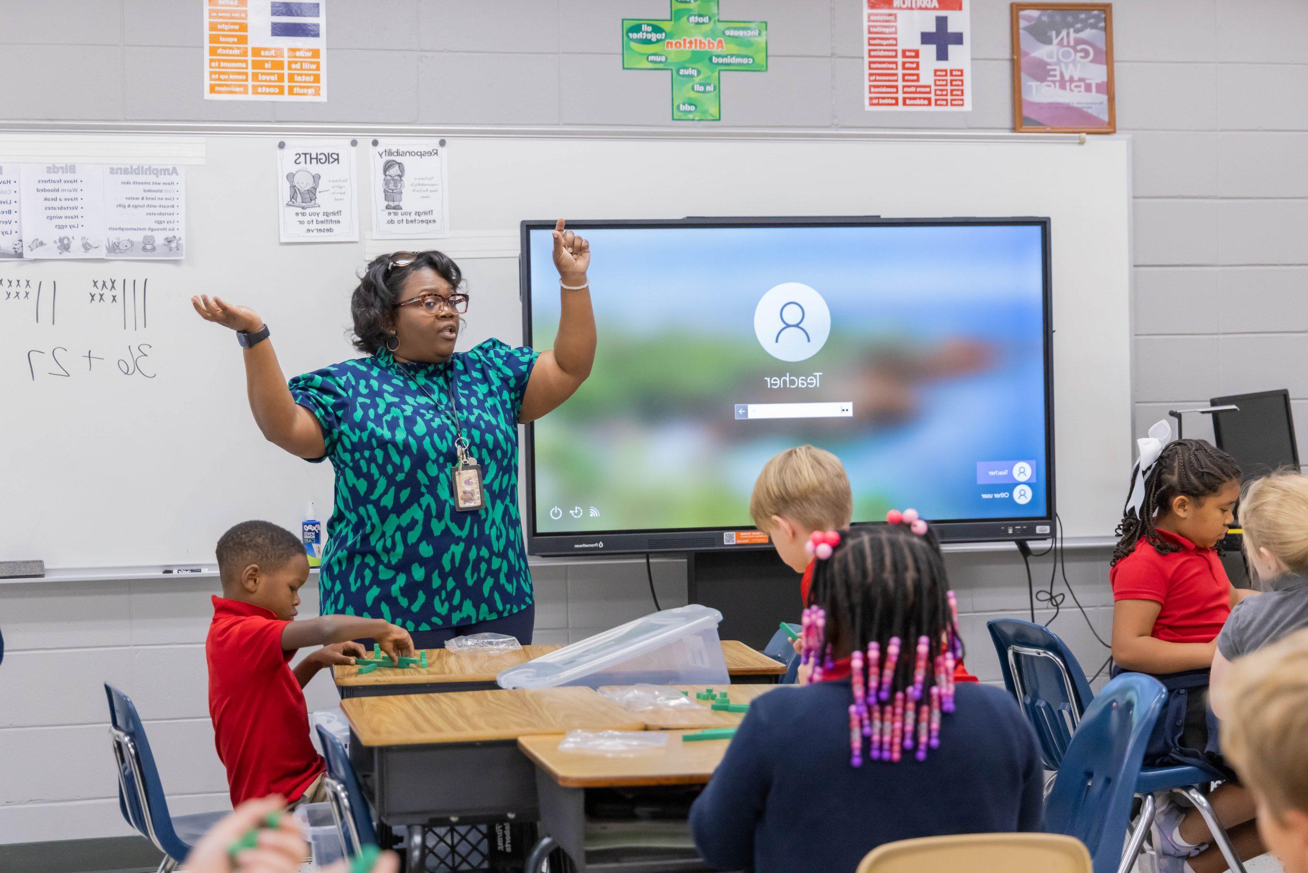 A teacher standing in front of elementary students.in a classroom with arms raised and students conducting activity at desks.